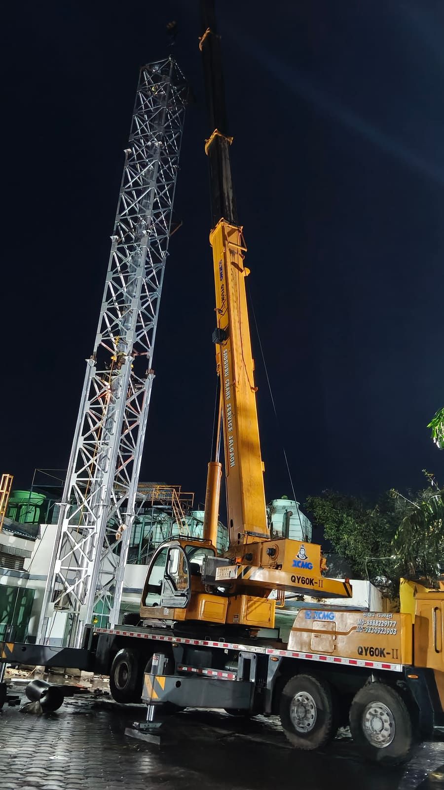 A galvanised lattice stack column held upright by a mobile crane during a night erection